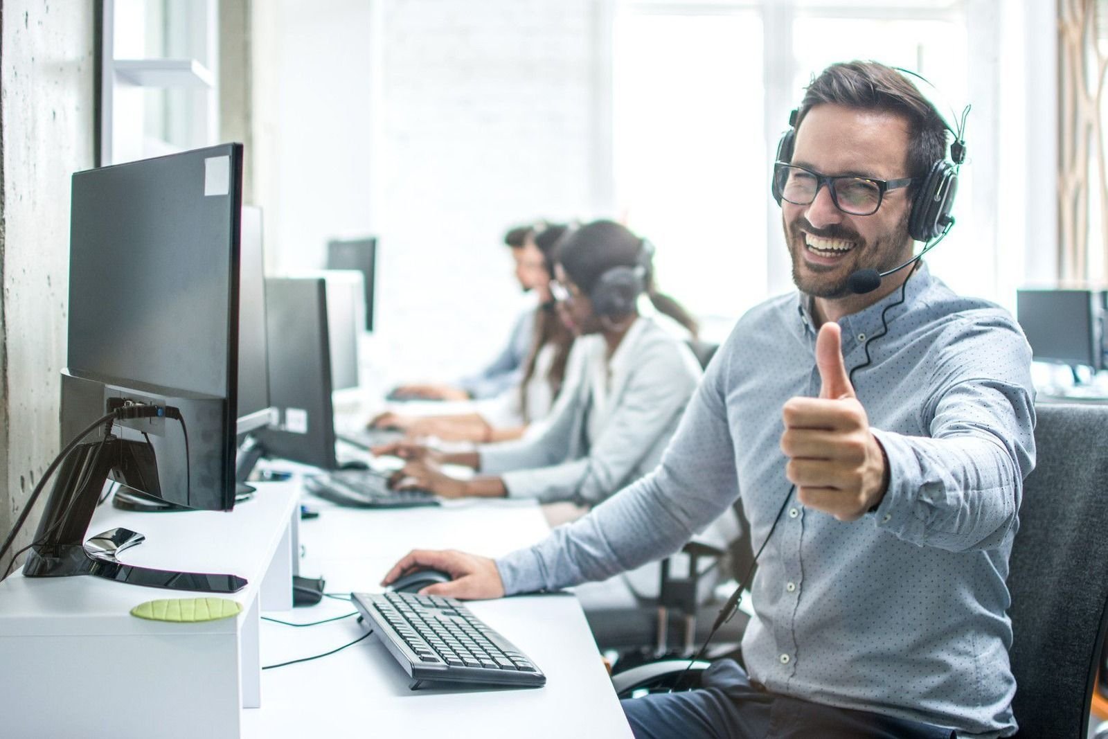 Smiling help desk technician with a headset giving a thumbs up, with support team members at their workstations in the background